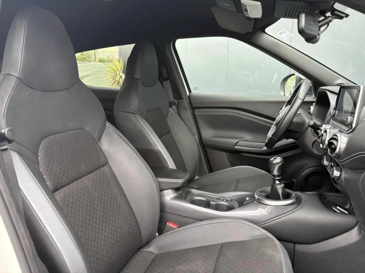 Intérieur du Nissan Juke 2024 vu côté conducteur, sièges noirs en TEP/tissu et levier de vitesse manuel visible.