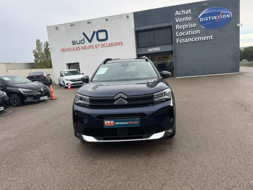 Citroën C5 Aircross hybride bleu foncé vue de face, avec barres de toit et feux allumés sur parking de concession.