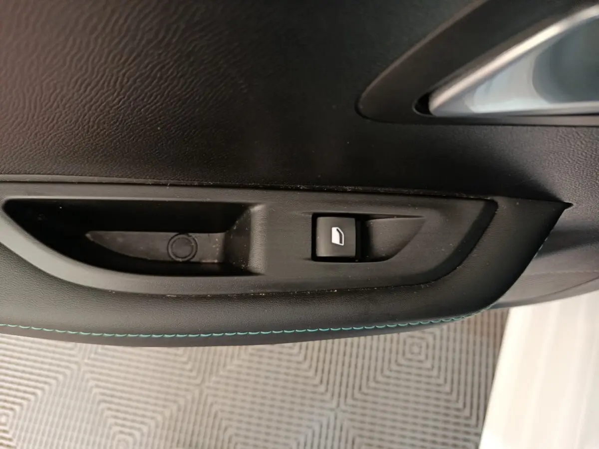 Détail de la commande de lève-vitre sur la porte intérieure noire avec surpiqûres bleues d’une Peugeot 208 blanche.