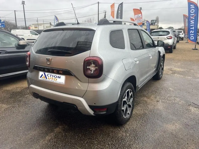 Vue 3/4 arrière droite d'un Dacia Duster gris Comète métallisé avec barres de toit et jantes alu distinctives.