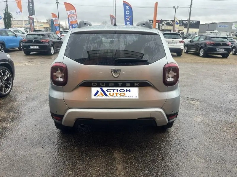 Vue arrière d'un Dacia Duster gris Comète métallisé avec logo et feux caractéristiques dans un parking humide.