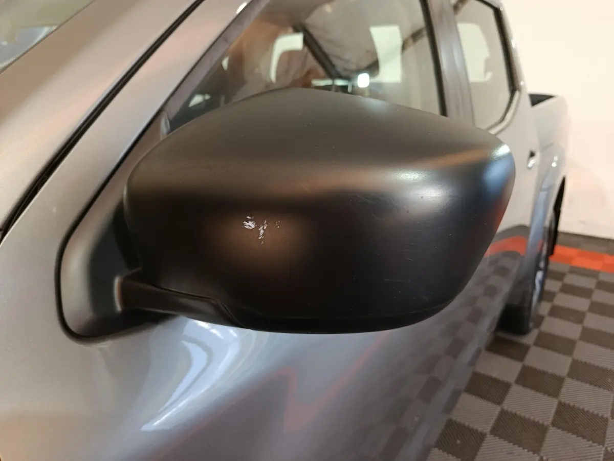 Gros plan sur le rétroviseur noir côté conducteur du Nissan Navara gris clair, vue partielle du profil avant.