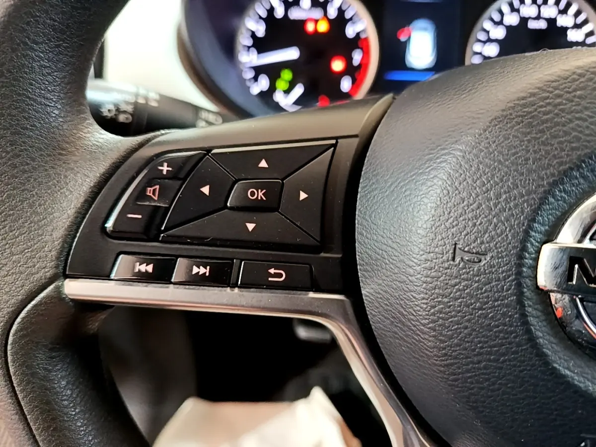 Gros plan sur le volant multifonctions noir de la Nissan Micra IG-T 100 Business Edition avec commandes audio et tableau de bord flou.