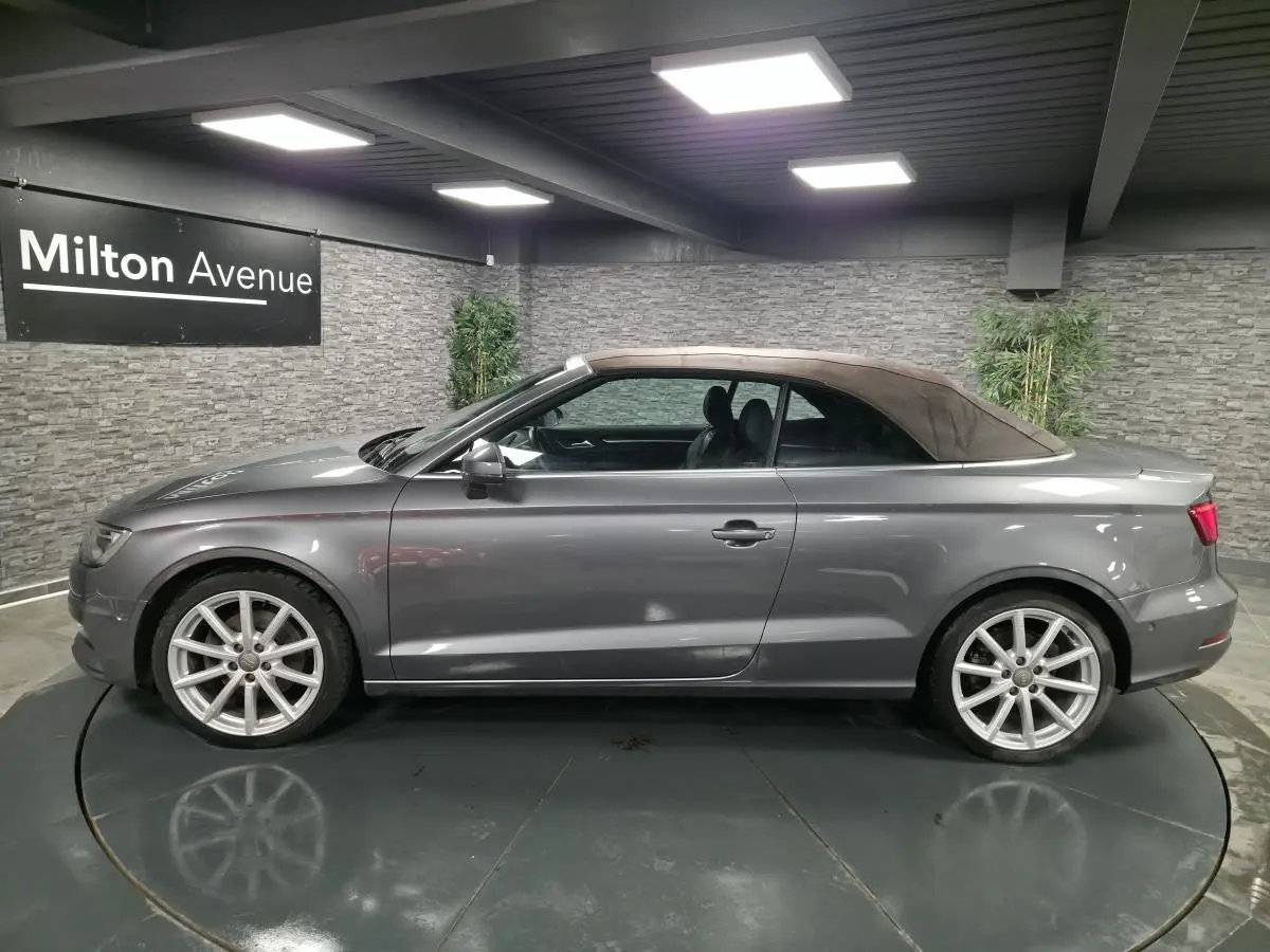 Profil côté gauche d'un Audi A3 Cabriolet gris foncé avec capote tissu marron dans un showroom.