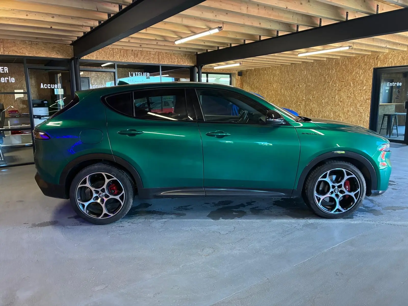 Vue de profil côté gauche d'une Alfa Romeo Tonale vert foncé avec jantes sport et freins rouges, stationnée en intérieur.