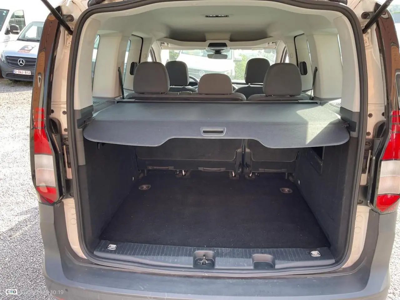 Vue arrière du coffre ouvert d'un Volkswagen Caddy blanc Candy 2024 avec cache-coffre enroulable et sièges arrière visibles.