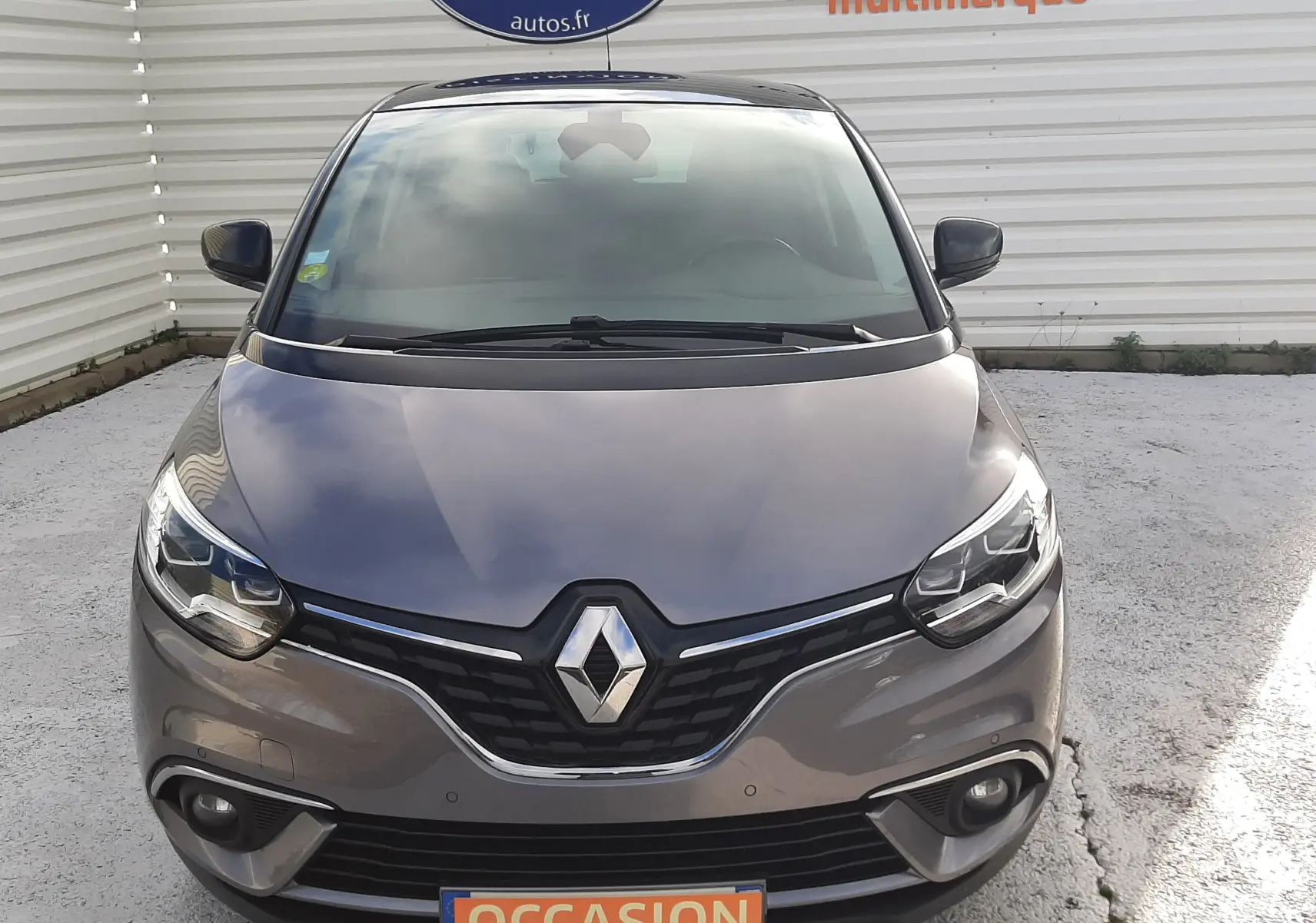 Vue avant d'une Renault Scénic IV gris Cassiopée avec toit noir, affichant une plaque "OCCASION" devant un mur blanc.