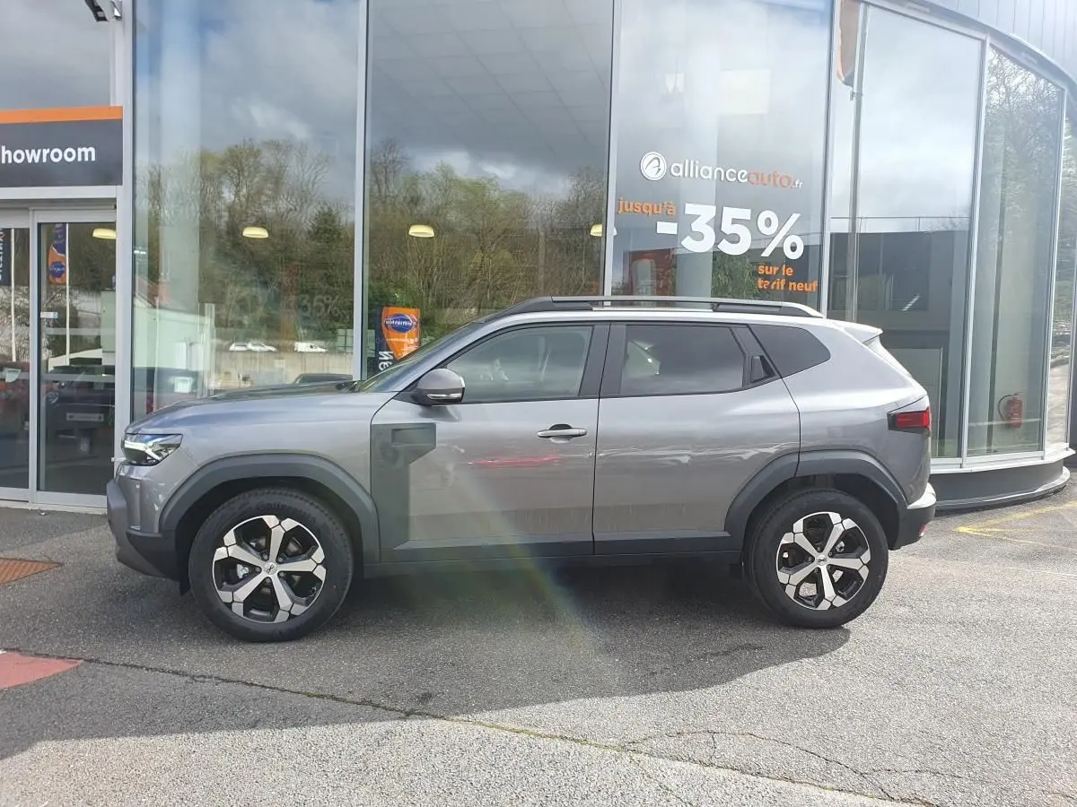 Vue de profil côté gauche d'un SUV Dacia gris schiste avec jantes alliage et barres de toit devant un showroom.