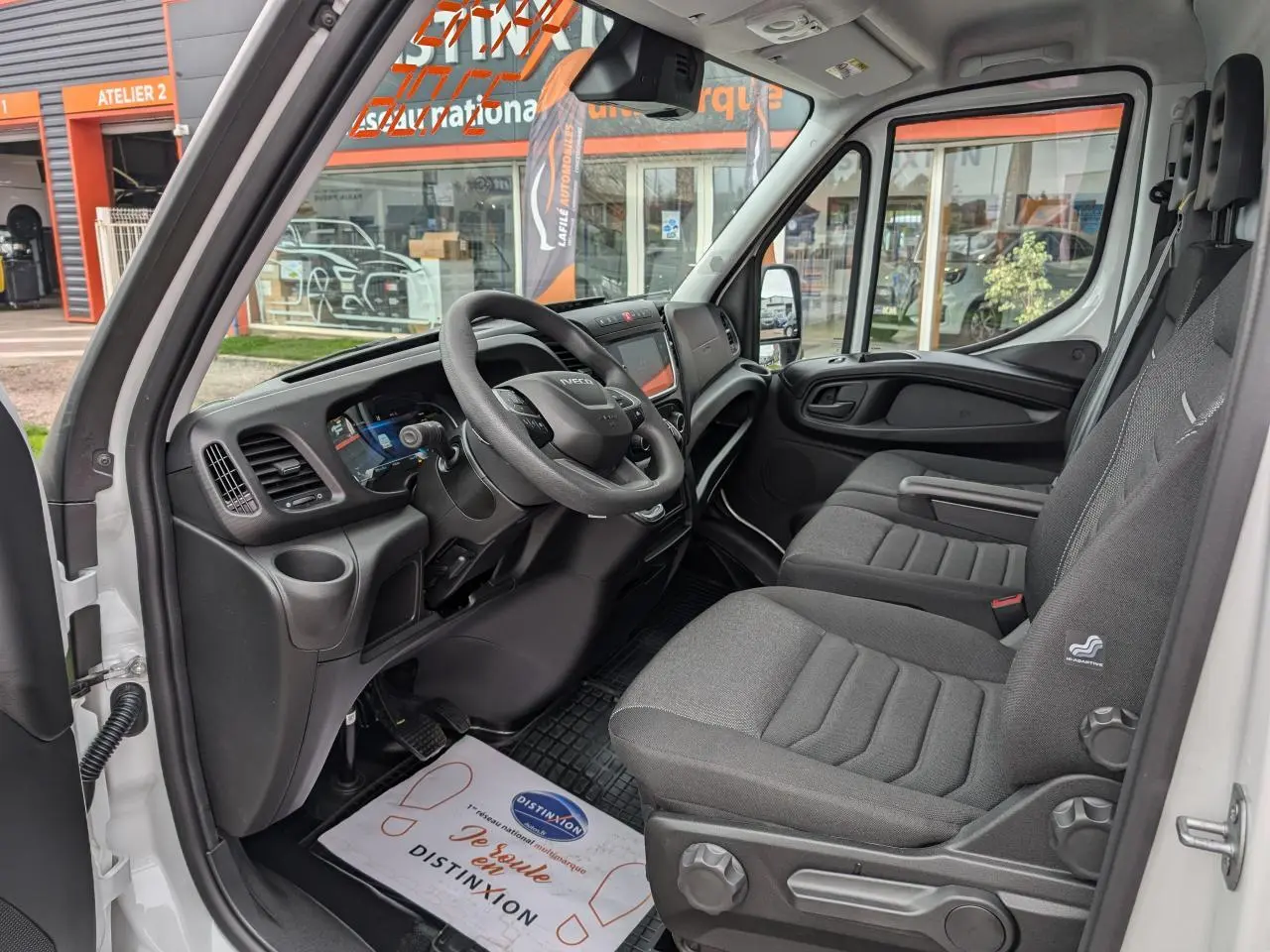Vue intérieure côté conducteur de l'IVECO Daily blanc 2025, montrant tableau de bord moderne et sièges en tissu gris.
