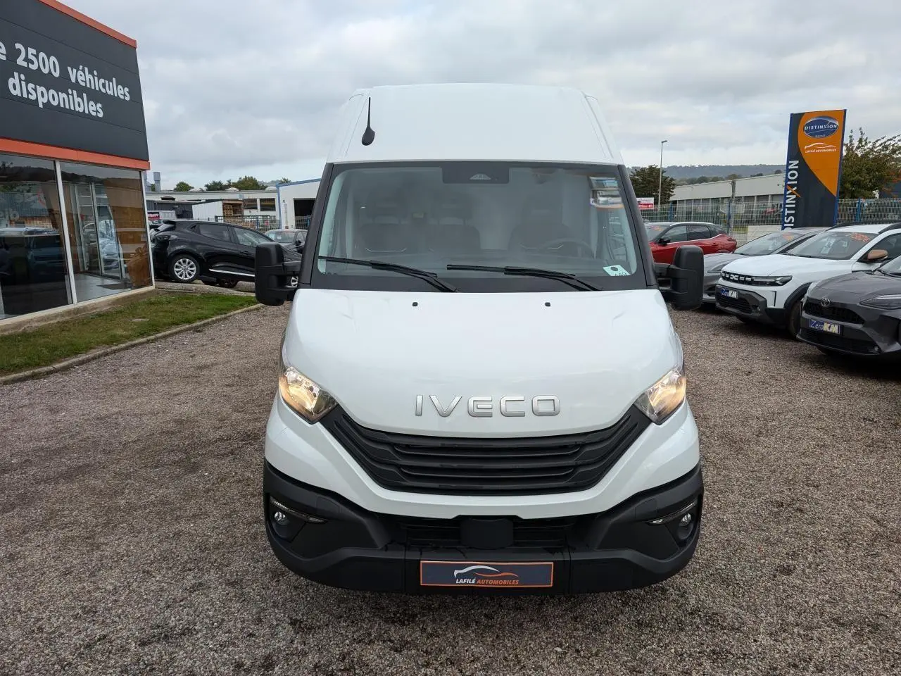 Fourgon Iveco Daily blanc vu de face, avec calandre noire et phares allumés sur un parking extérieur.