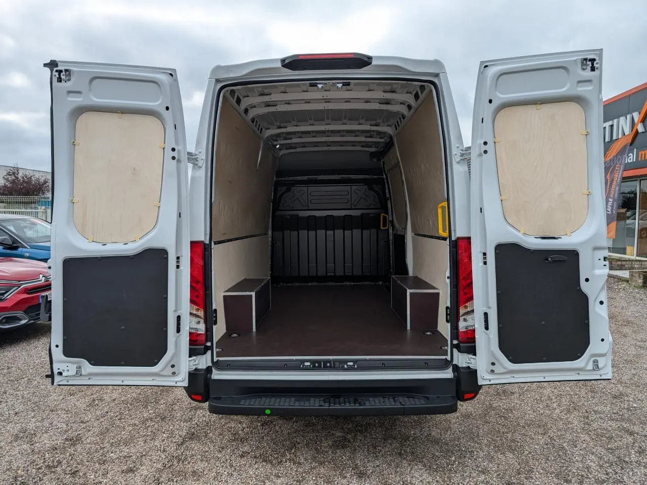 Vue arrière d’un utilitaire Iveco Daily blanc 2025 avec portes ouvertes et intérieur de chargement protégé par habillages bois et plancher antidérapant.