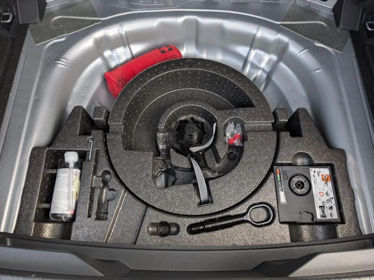 Vue plongeante sur le coffre ouvert d’un Volkswagen T-Roc gris, montrant la roue de secours et les outils de bord.