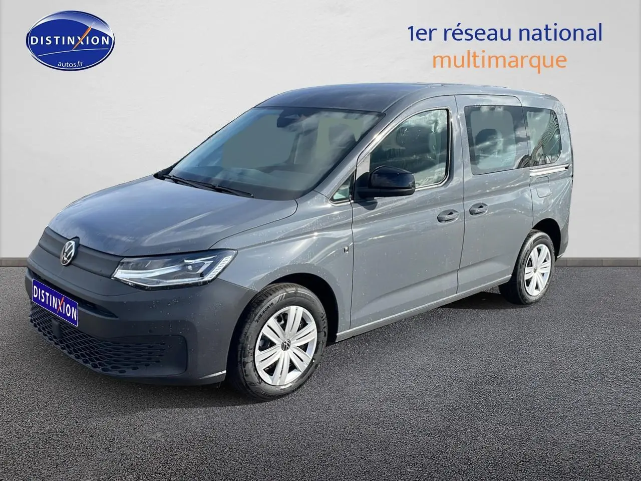 Volkswagen Caddy gris pur en 3/4 avant droit, avec barres de toit et jantes alliage visibles.