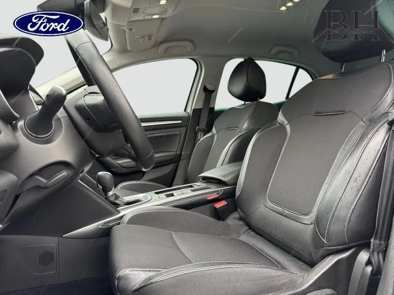 Vue intérieure côté conducteur montrant les sièges noirs en tissu et similicuir avec accoudoir central et console d'une Renault Mégane blanche.
