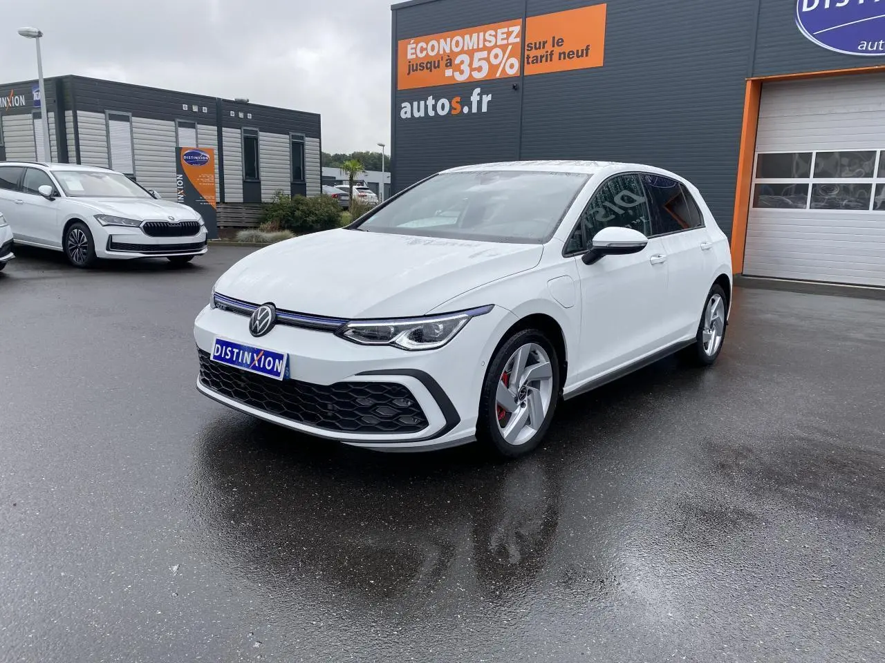 Volkswagen Golf 1.4 eHybrid blanche vue en 3/4 avant droit sur parking humide devant un bâtiment commercial.