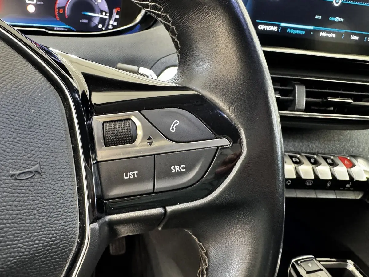 Gros plan sur le volant cuir noir de la Peugeot 5008 2022 avec commandes audio et tableau de bord numérique en arrière-plan.