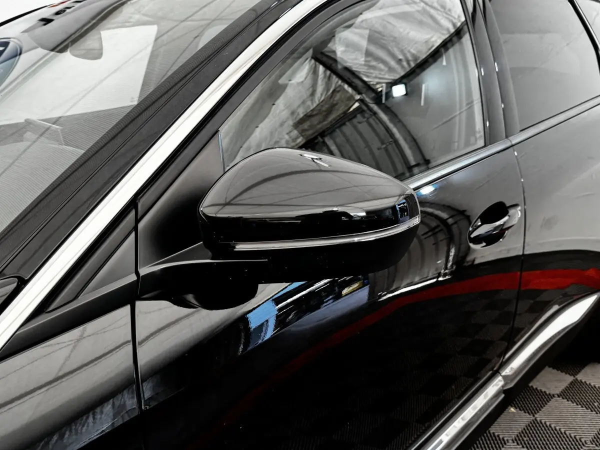Gros plan sur le rétroviseur côté conducteur noir brillant d’un Peugeot 5008 noir, avec reflet de l’environnement intérieur.