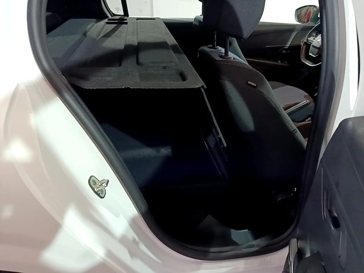 Vue intérieure depuis la porte arrière gauche d’une Peugeot 208 blanche, montrant le coffre avec tablette arrière relevée et siège avant noir.