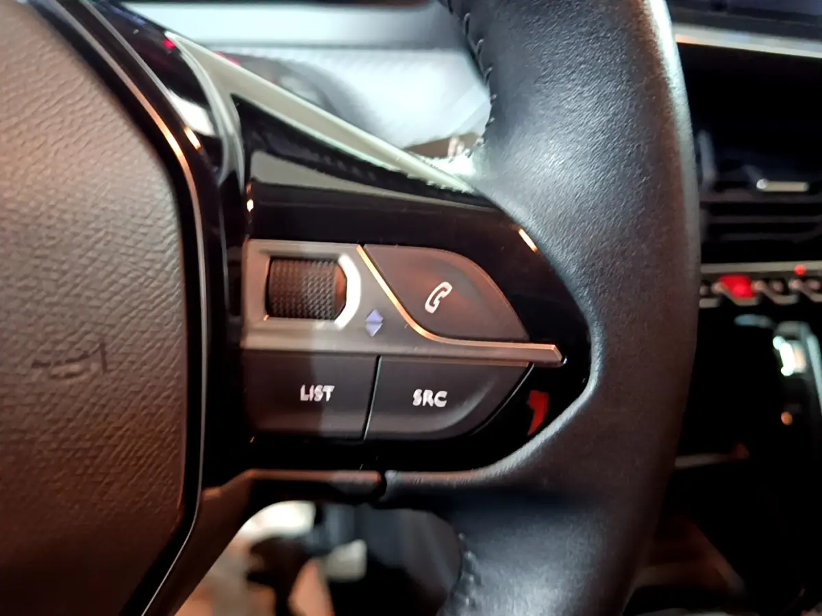 Gros plan sur les commandes gauche du volant noir de la Peugeot 208 1.5 BlueHDi 100 S&S Premium, avec boutons d’appel et réglages audio.