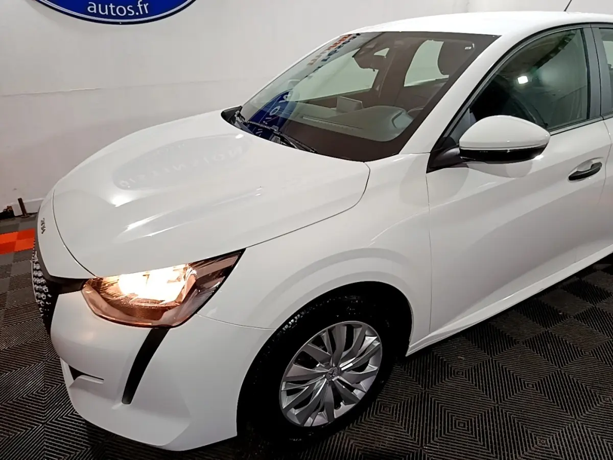 Vue 3/4 avant droit d'une Peugeot 208 blanche avec phares allumés et jantes alliage visibles.