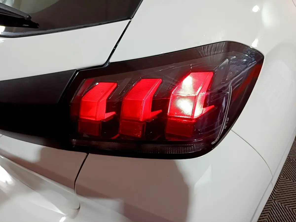 Gros plan sur le feu arrière droit rouge d’une Peugeot 208 blanche, soulignant le design en trois griffes distinctives.