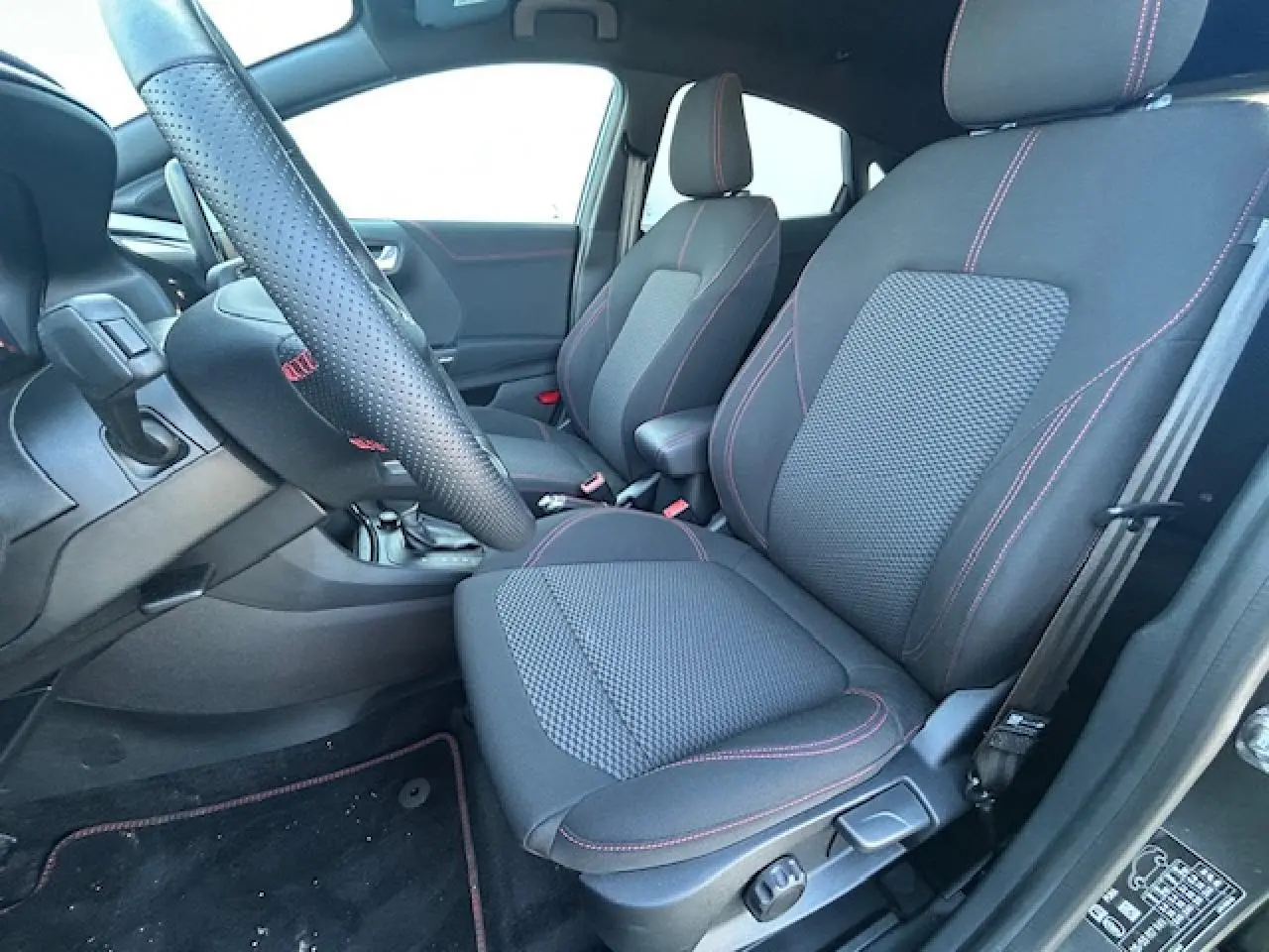 Vue intérieure côté conducteur du Ford Puma gris Magnetic 2023, sièges noirs avec surpiqûres rouges et volant perforé.