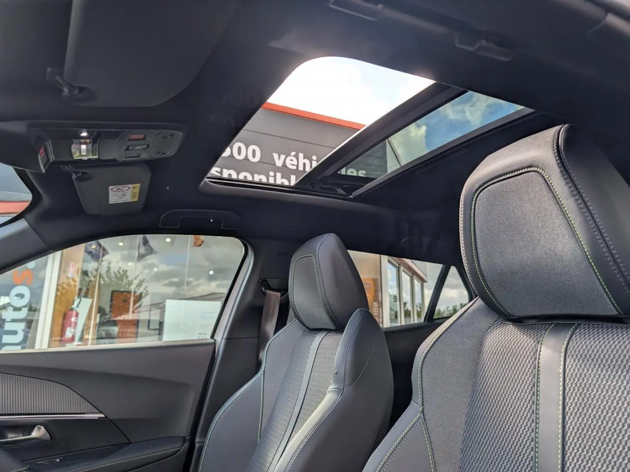Vue intérieure avant du Peugeot 2008 GT 2026 avec toit ouvrant panoramique et sièges tissu gris à surpiqûres vertes.