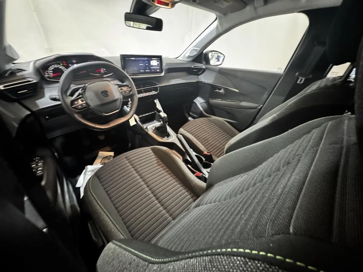 Vue intérieure côté conducteur de la Peugeot 208 2024, montrant le volant cuir, tableau de bord digital et sièges tissu gris.