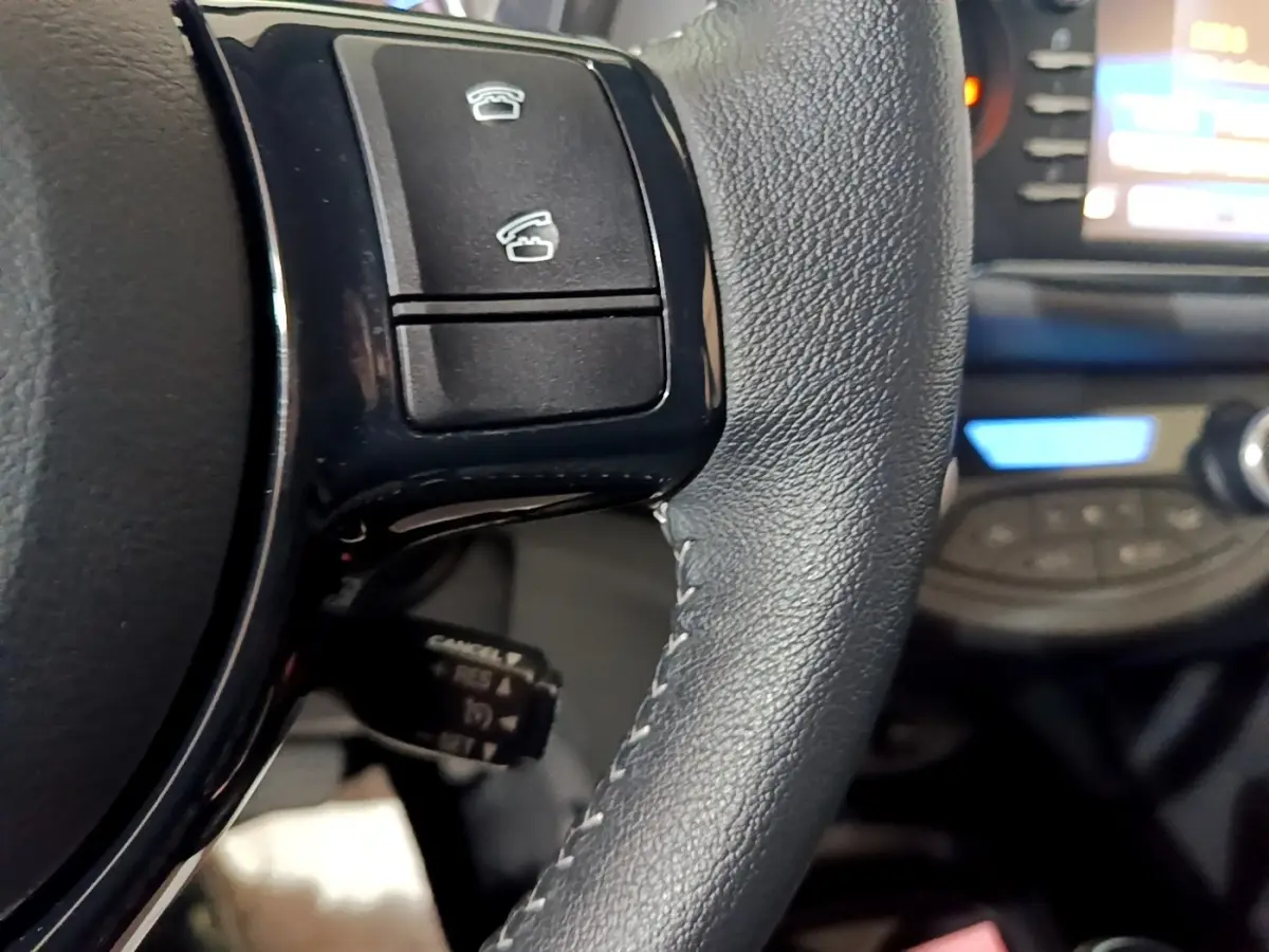 Gros plan sur le volant en cuir noir de la Toyota Yaris hybride 2019, avec commandes intégrées et tableau de bord flou en arrière-plan.