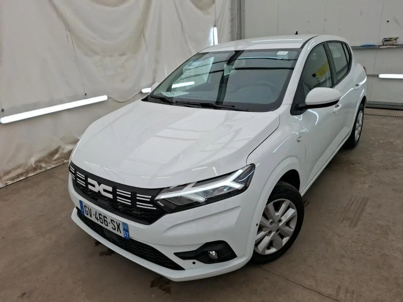 Vue 3/4 avant droit d'une Dacia Sandero blanche 2024 avec calandre noire et feux LED allumés dans un garage.