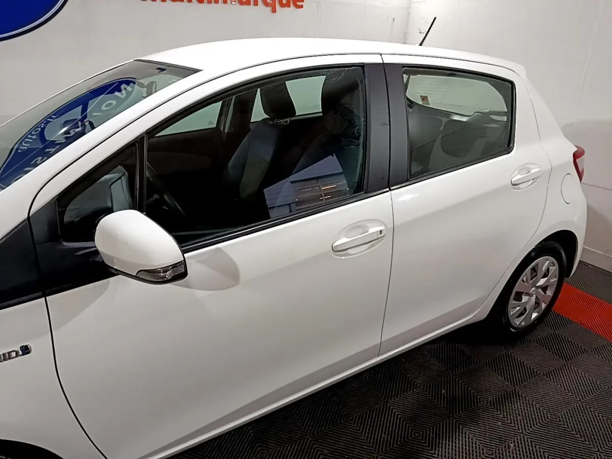 Profil côté gauche d'une Toyota Yaris blanche hybride 2019 en intérieur, mettant en valeur ses lignes compactes et ses rétroviseurs intégrés.