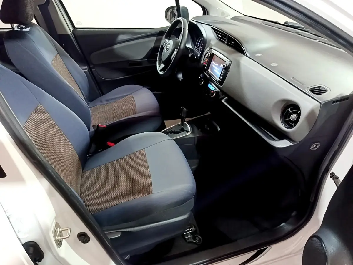 Vue intérieure côté conducteur de la Toyota Yaris blanche 2019, sièges tissu bicolore et tableau de bord avec écran tactile.