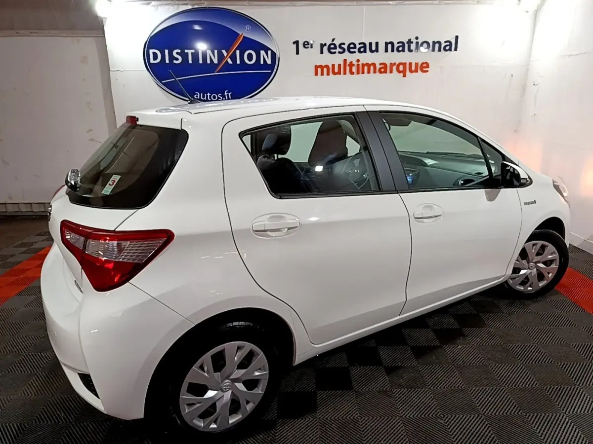 Vue 3/4 arrière droite d'une Toyota Yaris blanche 2019 hybride stationnée en intérieur sur sol damier noir et rouge.