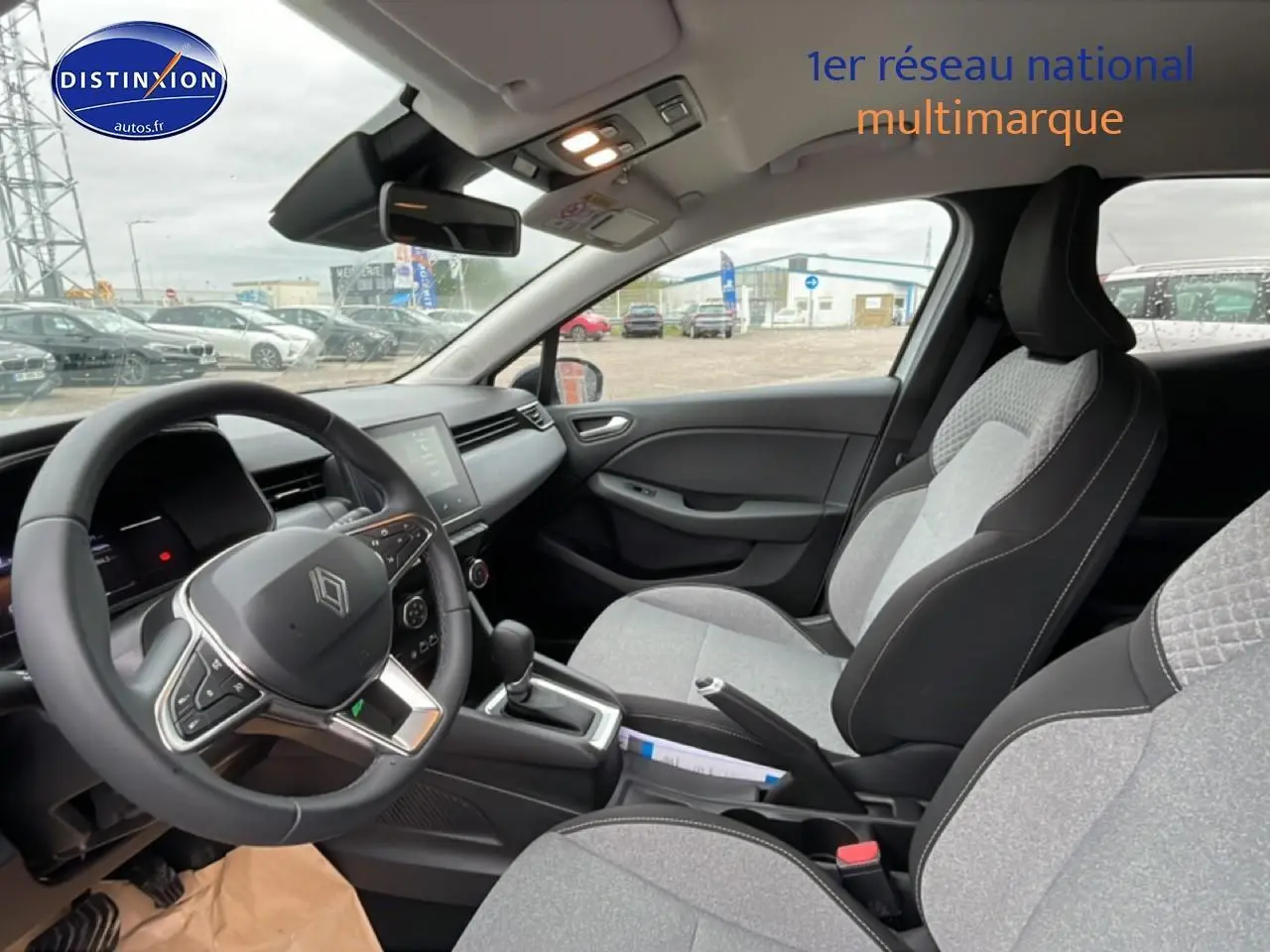 Vue intérieure côté conducteur de la Renault Clio V blanche, avec sièges gris à motifs 3D et tableau de bord moderne.