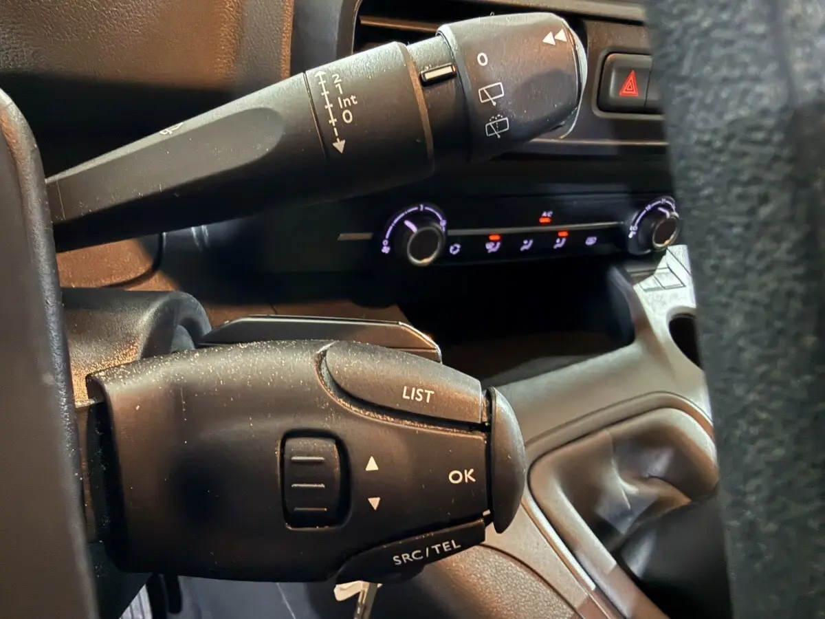 Gros plan sur les commandes au volant noir du Peugeot Rifter blanc, avec tableau de bord et commandes climatisation en arrière-plan.