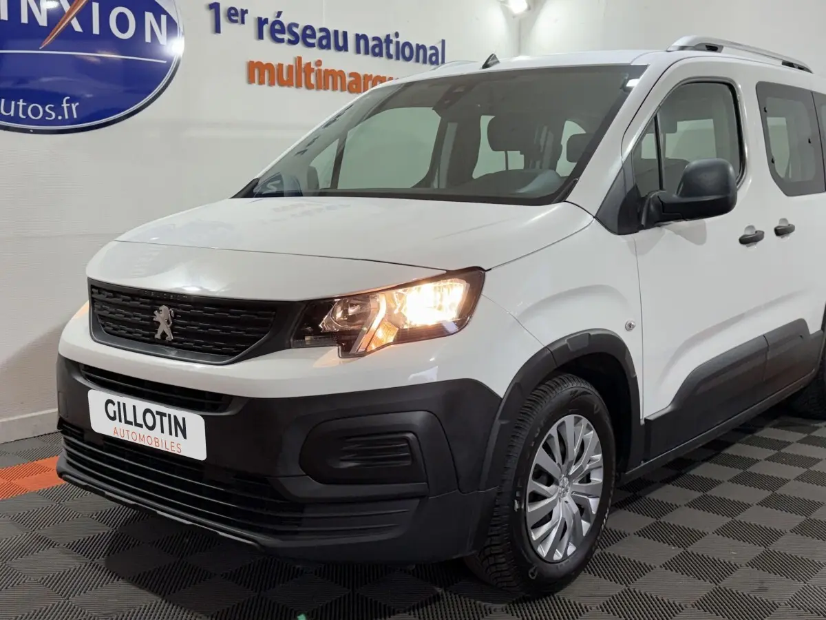 Vue 3/4 avant droit d’un Peugeot Rifter blanc de 2019 avec phares allumés et protections noires sur les bas de caisse.