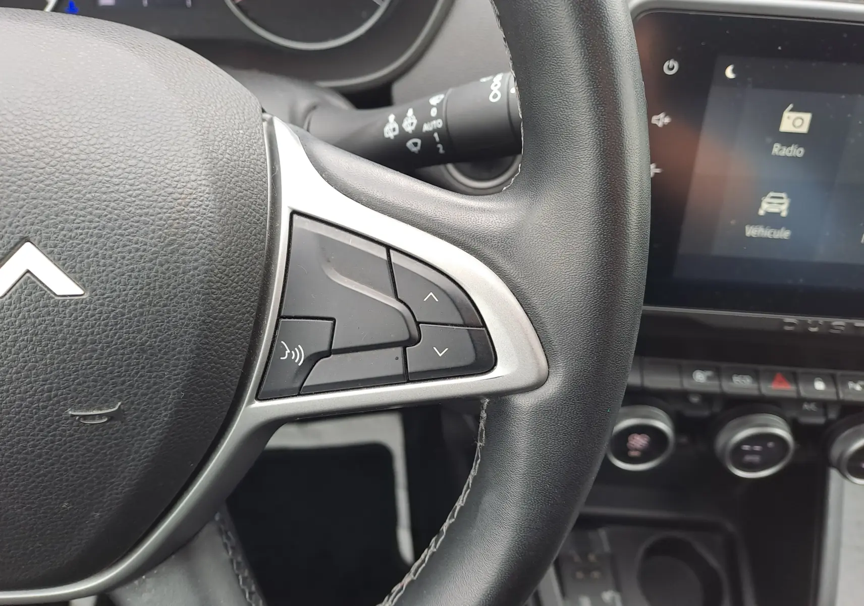 Gros plan sur le volant noir et les commandes intégrées, avec l’écran tactile et la console centrale du Dacia Duster 2024 en arrière-plan.