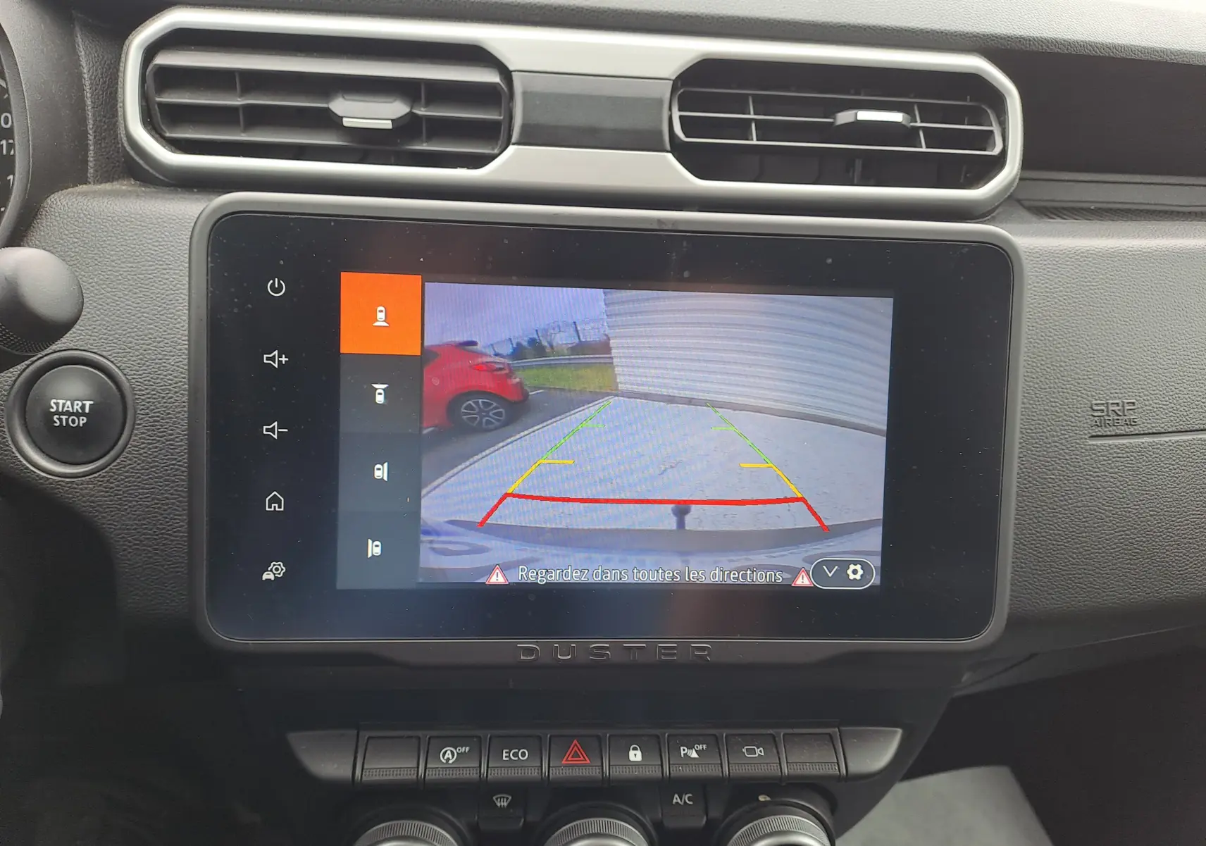 Écran central du Dacia Duster 2024 montrant la caméra de recul avec vue arrière et voiture rouge à gauche.
