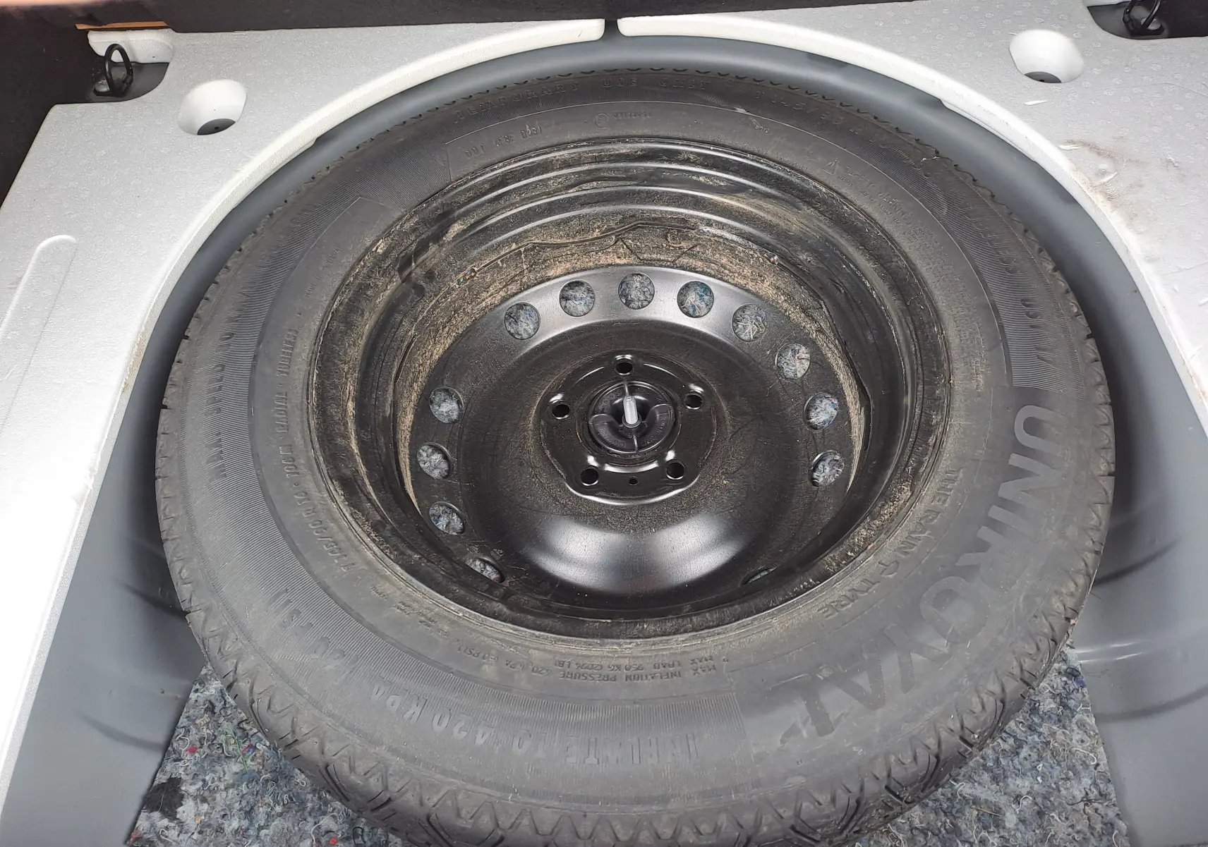 Roue de secours noire dans le coffre d'un Dacia Duster gris urbain, vue en plongée sur le pneu.