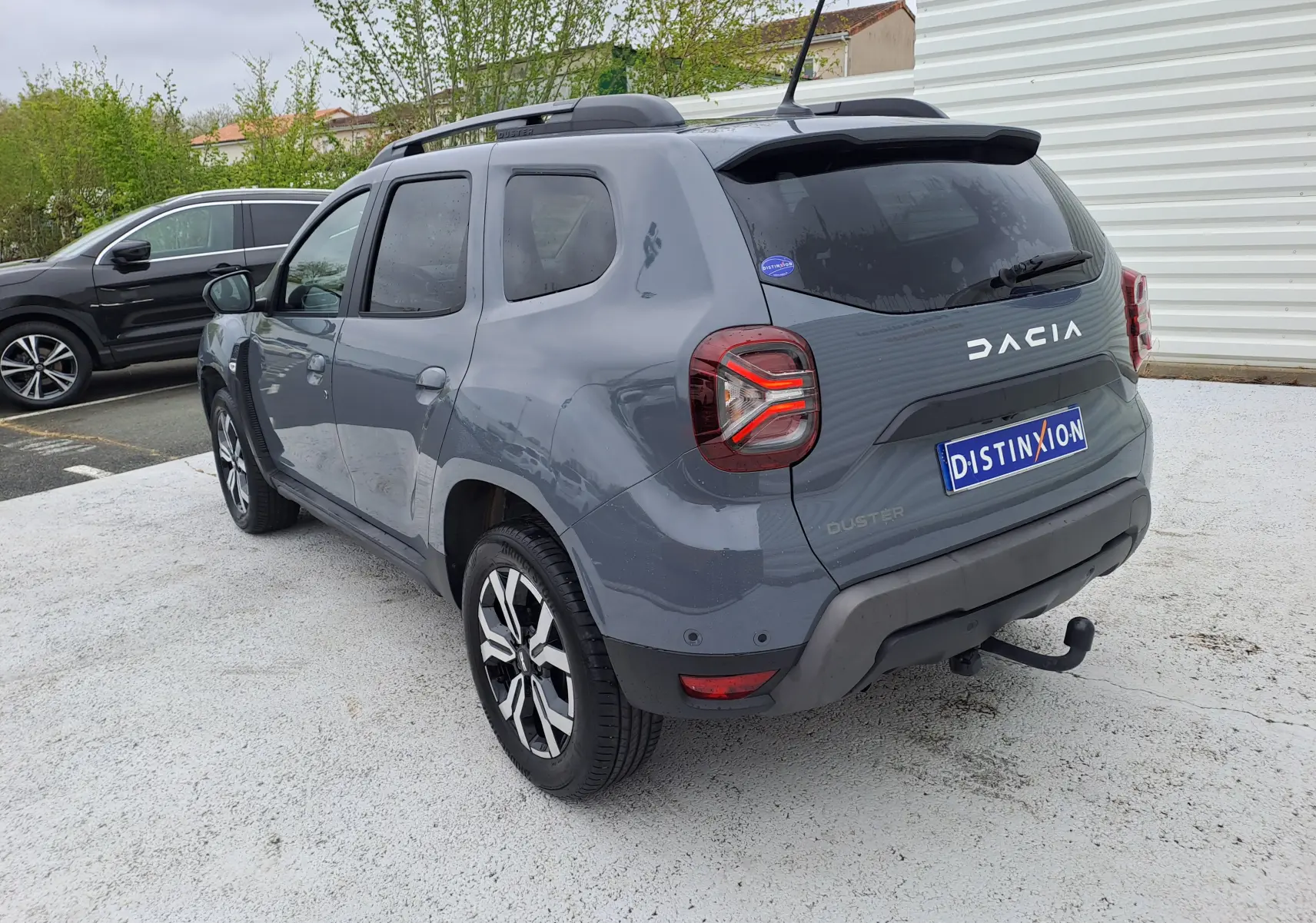 Vue 3/4 arrière droite d'un Dacia Duster gris urbain 2024 avec barres de toit et attelage visible.