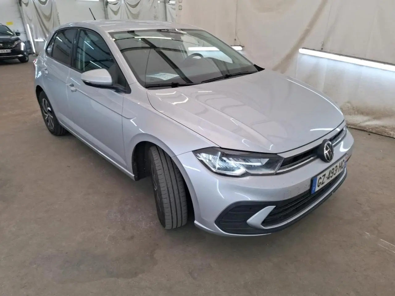 Volkswagen Polo gris clair vue 3/4 avant droit, avec calandre chromée et phares LED allumés.