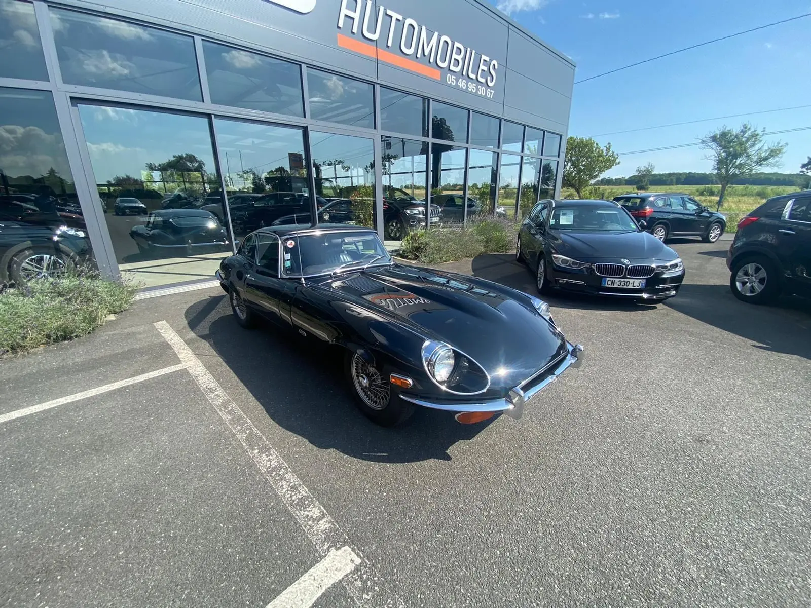 Jaguar Type E Coupé 4.2 Série 2 noire vue en 3/4 avant droit sur parking devant un showroom automobile.