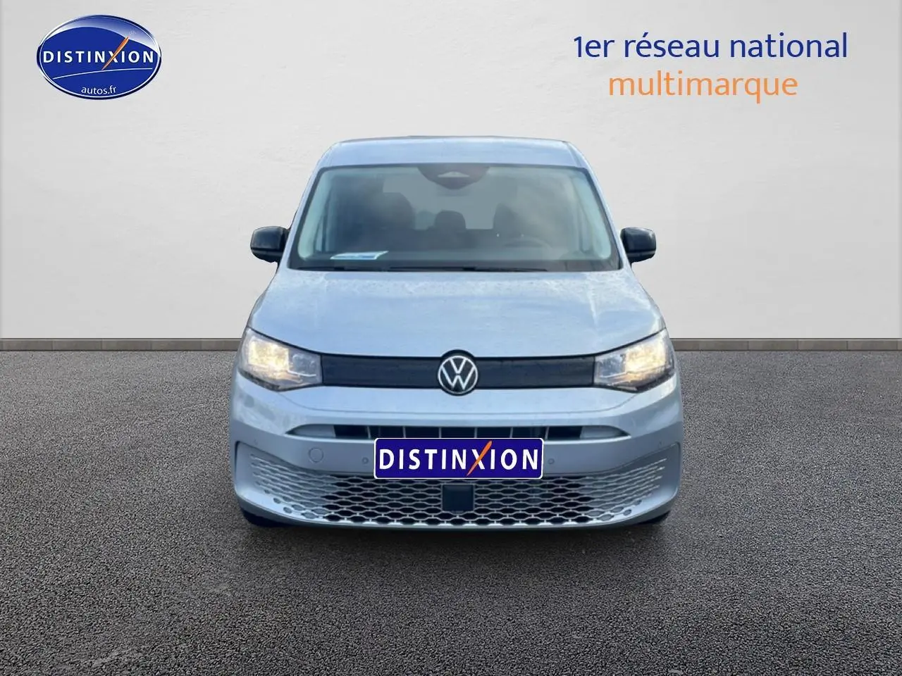 Vue de face d'un Volkswagen Caddy gris argent 2025 avec calandre noire et phares allumés.