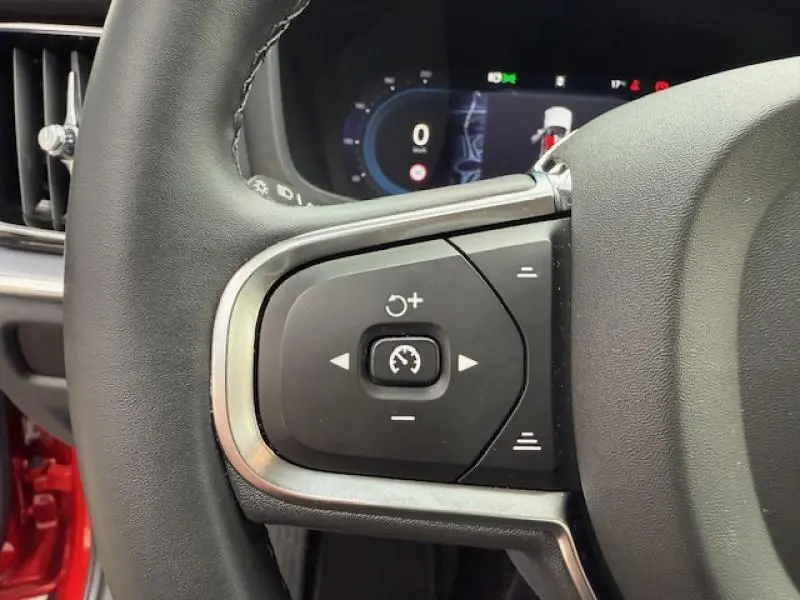Gros plan sur les commandes au volant gauche d'une Volvo S60 2024 rouge fusion métallisé, affichage numérique en arrière-plan.