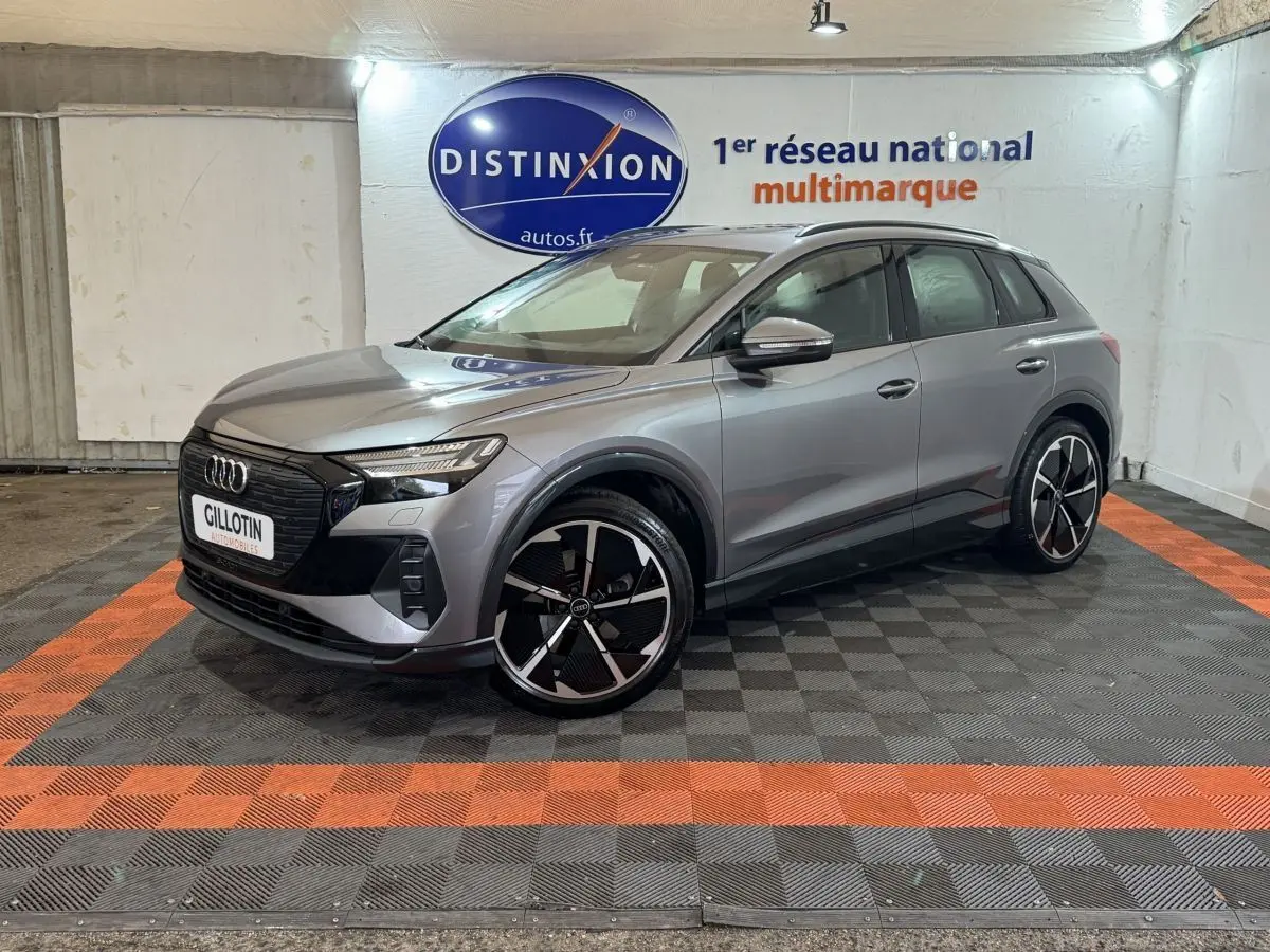 Audi Q4 E-TRON gris foncé en 3/4 avant droit, avec jantes noires et détails noirs brillants visibles en intérieur showroom.