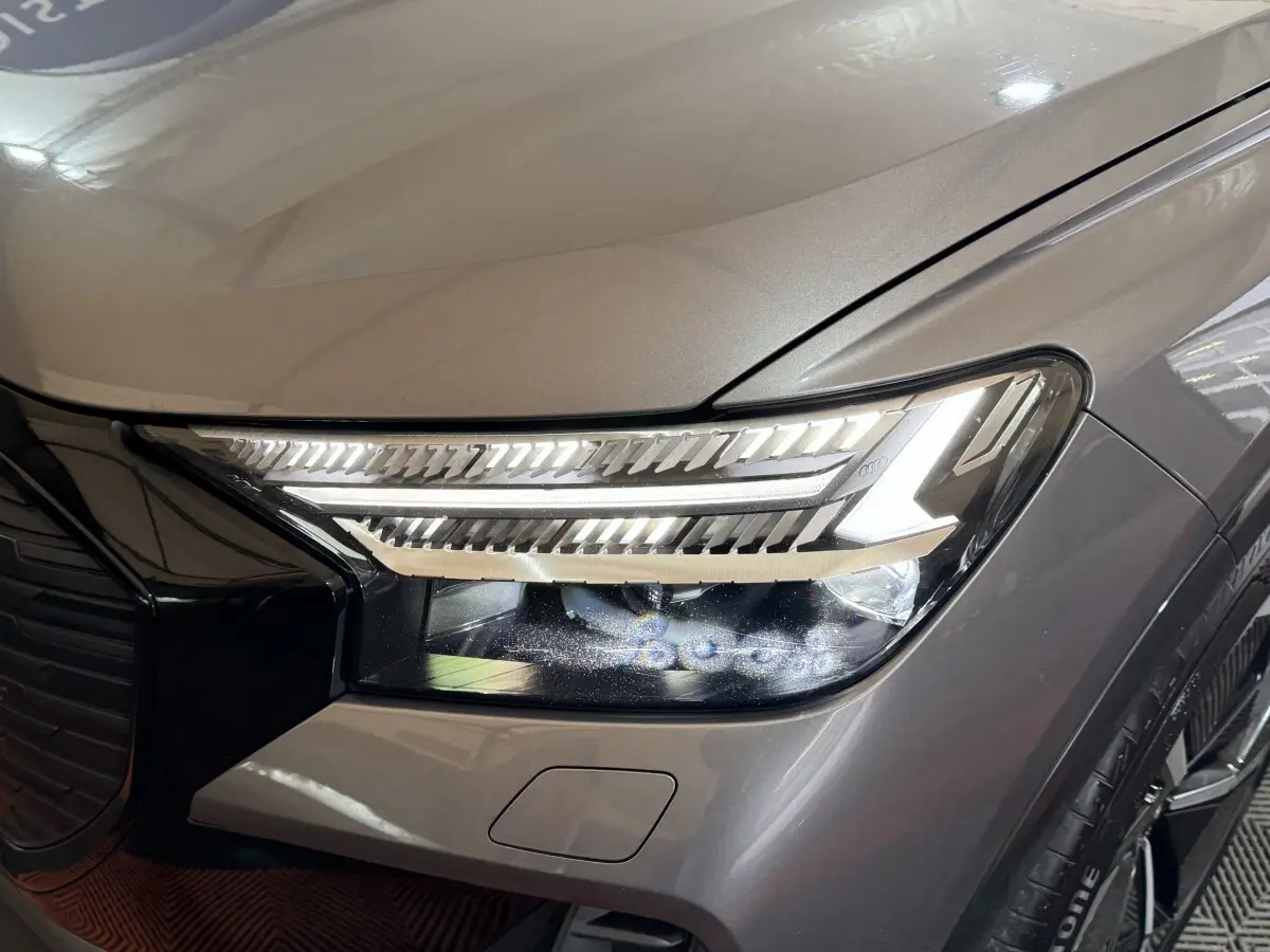 Gros plan sur le phare avant droit allumé d’un Audi Q4 E-TRON gris foncé avec détails LED et calandre noire brillante.