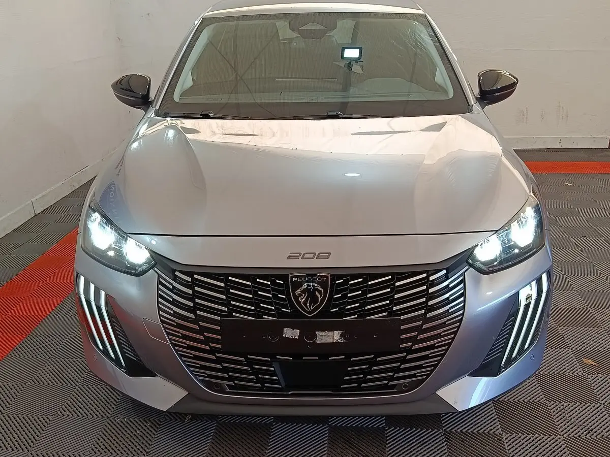 Vue avant d'une Peugeot 208 PureTech 100 S&S Allure gris clair avec phares LED et calandre noire distinctive allumés.