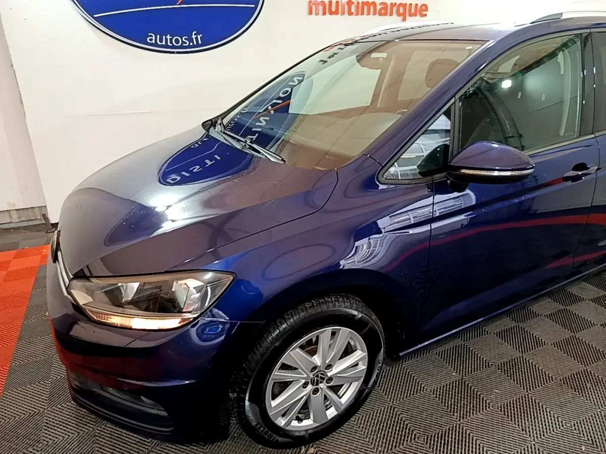 Vue 3/4 avant gauche d'un Volkswagen Touran bleu 2021 avec phares allumés et jantes alliage argentées.