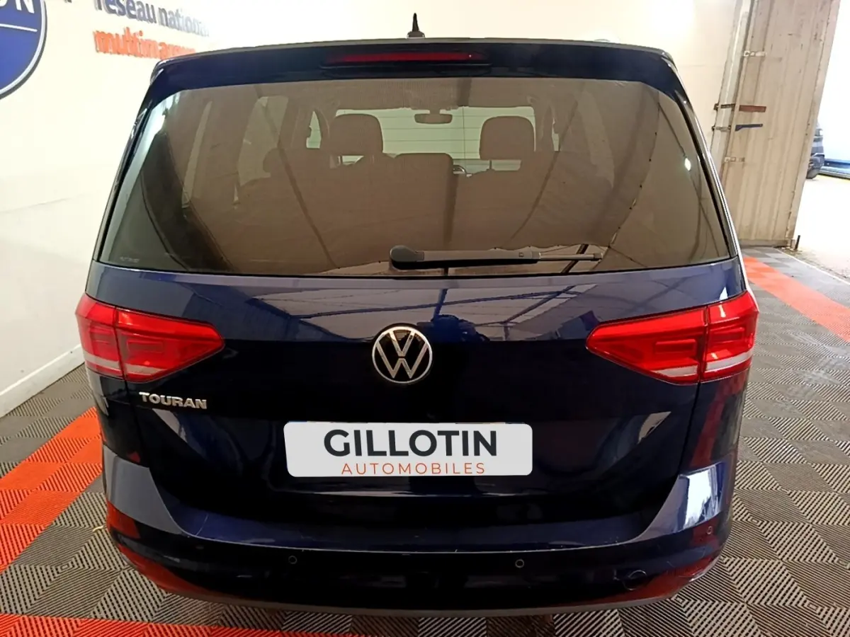Vue arrière d'un Volkswagen Touran bleu foncé garé en intérieur avec logo et feux rouges visibles.
