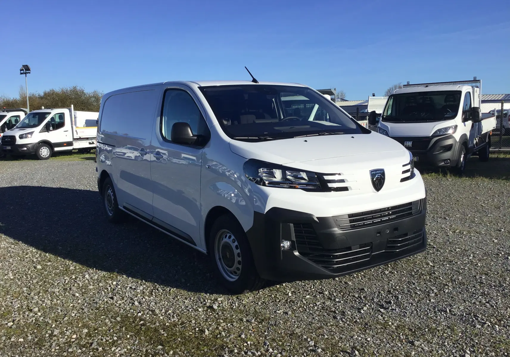 Fourgon Peugeot Expert blanc vu de 3/4 avant droit sur un parking extérieur avec d'autres véhicules utilitaires.